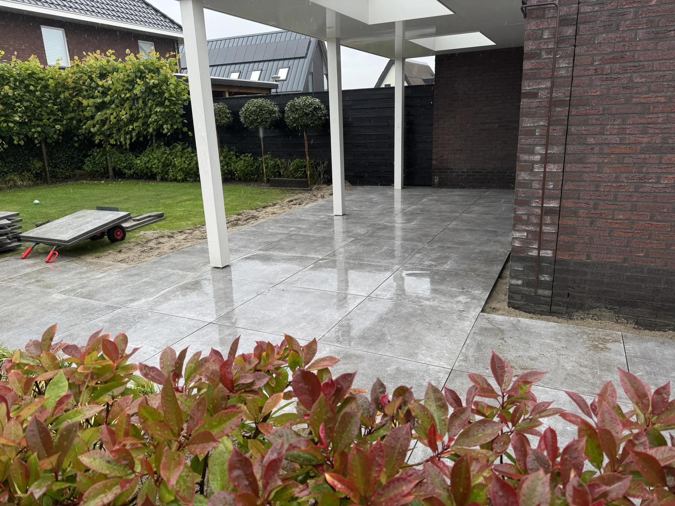 Terras van grote keramische tegels onder een overkapping, met een strakke kantopsluiting langs het gazon